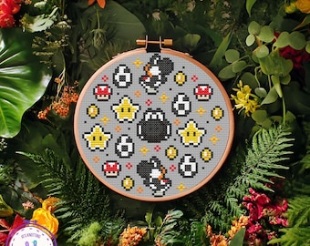 May include: A cross-stitch embroidery hoop featuring a gray background with pixelated images of a black Yoshi, yellow stars, red mushrooms, and coins. The embroidery is set in a wooden hoop and surrounded by greenery and flowers.
