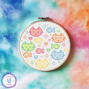 May include: A cross-stitch embroidery hoop with a design of pixelated cats and fish in various colours. The cats are pink, orange, green, blue, and yellow. The background is white, and the hoop is made of wood.