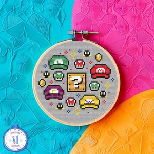May include: A cross-stitch embroidery hoop featuring a pixelated design of video game elements, including hats, coins, and a question mark block. The design is set against a gray fabric, with a wooden hoop. The background is a mix of bright colors.