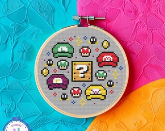 May include: A cross-stitch embroidery hoop featuring a pixelated design of video game elements, including hats, coins, and a question mark block. The design is set against a gray fabric, with a wooden hoop. The background is a mix of bright colors.