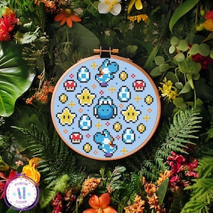 May include: A cross-stitch embroidery hoop featuring a light blue background with a pattern of Yoshi characters, stars, eggs, and mushrooms. The design is in pixelated style, with a wooden hoop. The background is a mix of green foliage and colorful flowers.