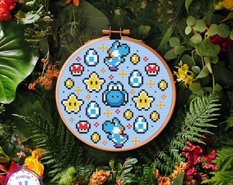 May include: A cross-stitch embroidery hoop featuring a light blue background with a pattern of Yoshi characters, stars, eggs, and mushrooms. The design is in pixelated style, with a wooden hoop. The background is a mix of green foliage and colorful flowers.