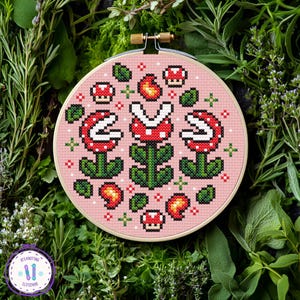 May include: A cross-stitch embroidery hoop featuring a pink background with a pixelated design of red and white piranha plants, mushrooms, and fire flowers. The embroidery is set in a wooden hoop, surrounded by green foliage.