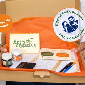Könnte beinhalten: Ein Bastelset in einem Karton, gefüllt mit orangefarbenem Seidenpapier und verschiedenen Materialien. Enthält Farbtöpfe, einen hölzernen Stickrahmen und gedruckte Materialien. Das Set trägt den Text "Let's get creative".