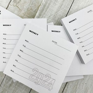 May include: A set of four white weekly planner notepads with black text and lines. The notepads have the word "WEEKLY" printed at the top and include the days of the week: Monday, Tuesday, Wednesday, Thursday, Friday, Saturday, and Sunday. One notepad has the year "2024" printed in a large, bold font.