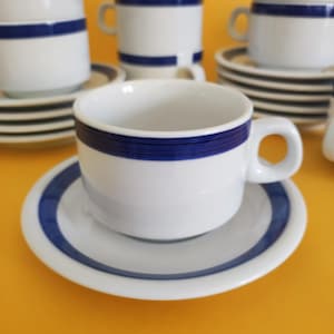 Op de afbeelding: Een set witte keramische koffiekopjes met blauwe strepen. De kopjes en schoteltjes zijn gestapeld op een geel oppervlak.