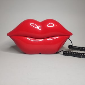 May include: A glossy, red telephone shaped like a pair of lips. The phone has a black, coiled cord. The design is a novelty item, adding a pop of color and a unique aesthetic to any space.
