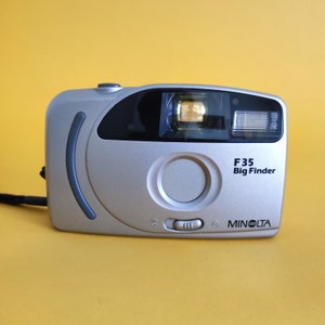 May include: A silver Minolta F35 Big Finder camera with a black lens and flash. The camera has a large circular lens and a black strap. The text "F35 Big Finder" and "Minolta" are printed on the front of the camera.