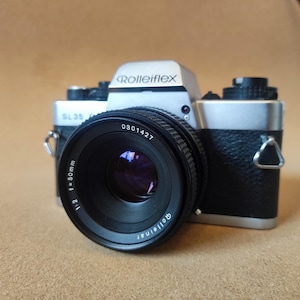 May include: A vintage Rolleiflex SL35 camera with a black and silver body and a prominent lens. The lens is marked with "f=50mm 1:2" and "Rolleinar." The camera's top has the brand name "Rolleiflex" in silver lettering.