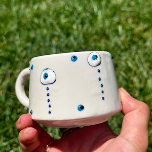 Könnte beinhalten: Weiße Keramiktasse mit blauen bösen Augen-Designs. Die Tasse hat einen Henkel und ist mit mehreren blauen Augen und blauen Punkten verziert.