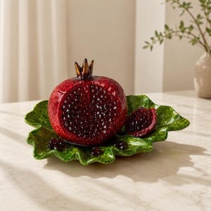 Puede incluir: Una granada decorativa de cerámica con una corona de bronce se encuentra sobre un plato verde con forma de hoja. La granada es de color rojo intenso con semillas visibles. Una rodaja de granada también está en el plato.