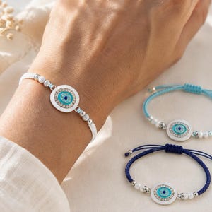 May include: Three adjustable bracelets featuring a central circular charm with a blue and white evil eye motif surrounded by small clear crystals. The bracelets are strung on white, light blue, and dark blue cord with silver-toned beads.