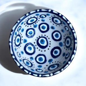 Może przedstawiać: Biała ceramiczna miska z niebiesko-białym wzorem złego oka. Miska jest ozdobiona powtarzającym się wzorem niebieskich i białych oczu, otoczonych małymi czarnymi kropkami.
