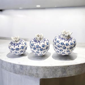 Może przedstawiać: Trzy białe ceramiczne wazony w kształcie granatu z niebieskimi i białymi wzorami złych oczu. Wazony są ułożone w rzędzie od najmniejszego do największego.