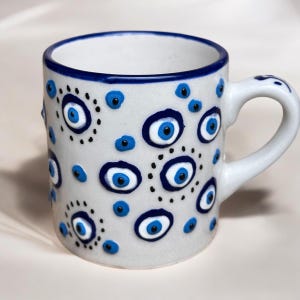 May include: White ceramic mug with a blue and white evil eye design. The mug has a handle and is decorated with multiple blue and white evil eyes.
