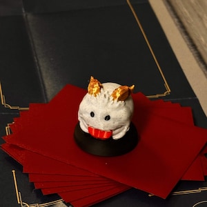 Pode incluir: Uma figura branca estilizada de criatura com detalhes dourados nas orelhas e boca vermelha, sobre uma base preta. Descansa sobre uma pilha de cartas vermelhas, com um tabuleiro de jogo azul escuro ao fundo.