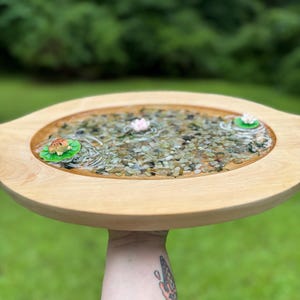 May include: A wooden bird bath with a resin pond design. The pond features small stones, a frog on a lily pad, and two small pink flowers. The bird bath is held up by a wooden base.