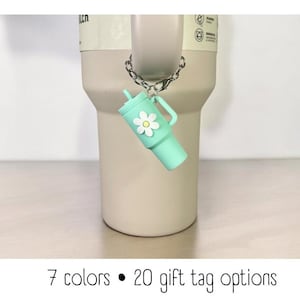 May include: A light green tumbler charm with a white daisy design hangs from a silver chain. The charm is attached to a light brown tumbler. The text "7 colors &bull; 20 gift tag options" is at the bottom of the image.