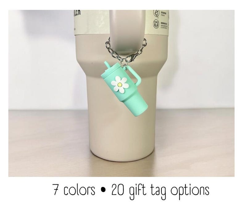 May include: A light green tumbler charm with a white daisy design hangs from a silver chain. The charm is attached to a light brown tumbler. The text "7 colors &bull; 20 gift tag options" is at the bottom of the image.