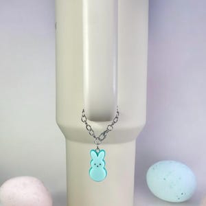 May include: A light blue bunny charm hangs from a silver chain attached to a white travel mug. The mug has a blue lid and a handle. The charm is a popular Easter candy.