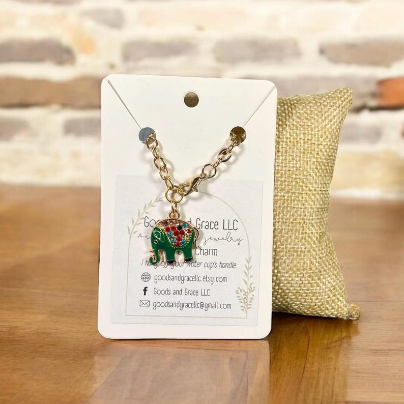 May include: A gold-colored elephant charm with red and green accents, attached to a gold chain. The charm is displayed on a white card with the text "Goods and Grace LLC jewelry charm". A burlap pillow is in the background.
