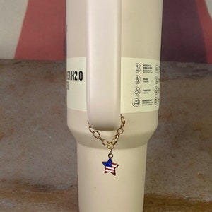 May include: A white tumbler with a gold chain and a star-shaped charm with the American flag design. The tumbler has a stainless steel lid and a handle.