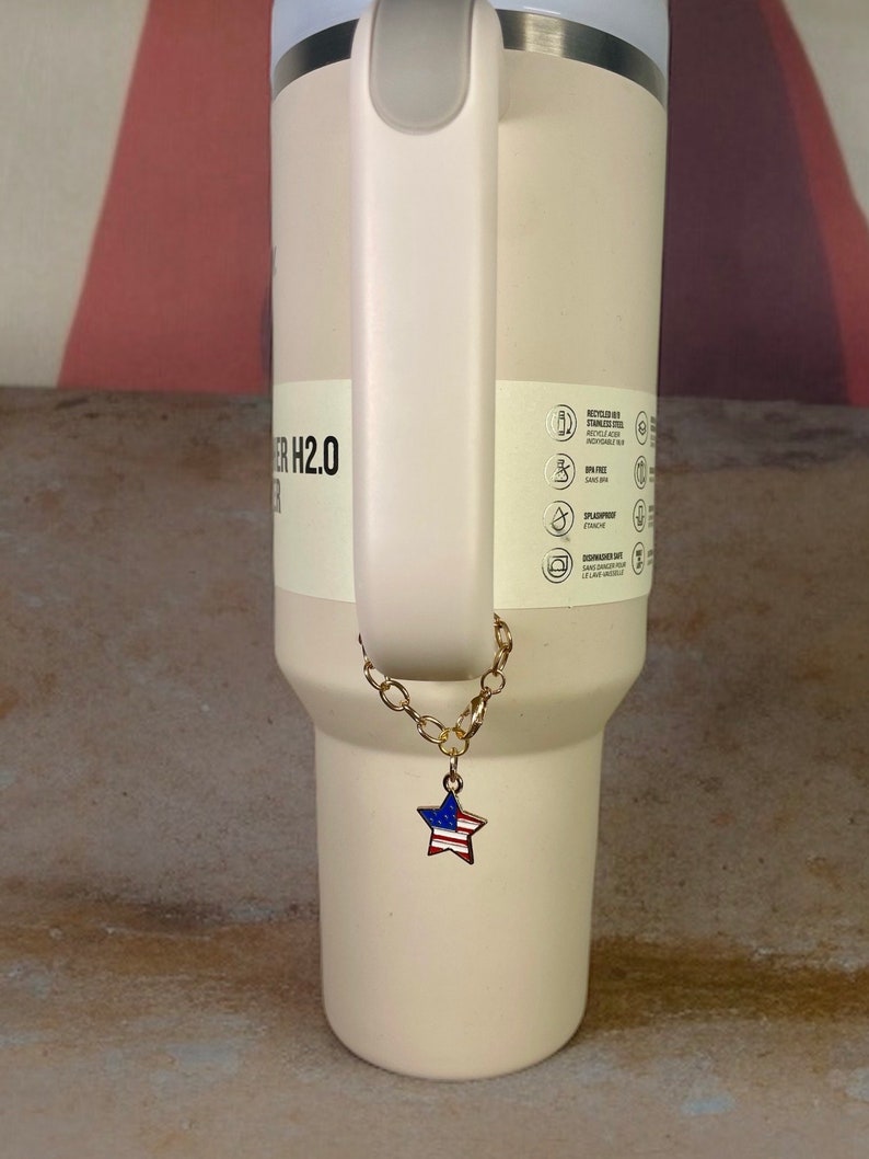 May include: A white tumbler with a gold chain and a star-shaped charm with the American flag design. The tumbler has a stainless steel lid and a handle.