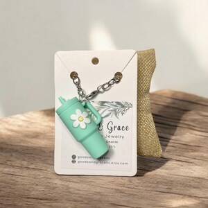 May include: A teal blue charm with a white daisy on it, attached to a silver chain. The charm is shaped like a cup and has the text "Good & Grace Jewelry Charm (25')" on a white card behind it.