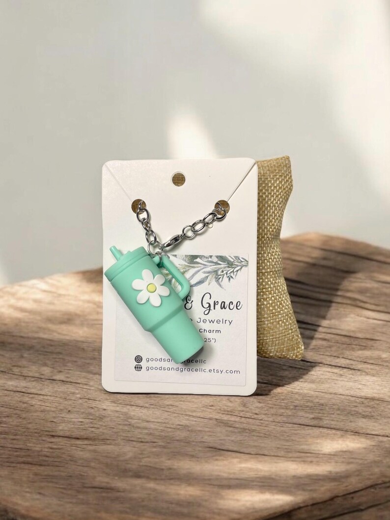 May include: A teal blue charm with a white daisy on it, attached to a silver chain. The charm is shaped like a cup and has the text "Good & Grace Jewelry Charm (25')" on a white card behind it.