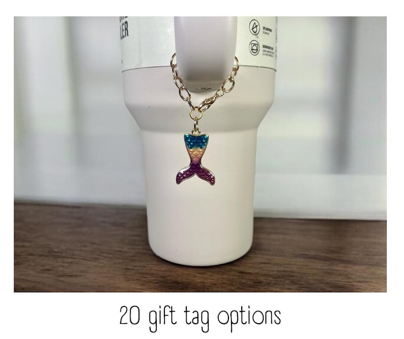 May include: A gold chain with a mermaid tail charm. The charm is iridescent with blue, green, and purple scales. The chain is attached to a white tumbler.