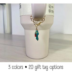 May include: A white ceramic mug with a gold chain and a blue and red bird charm. The mug has a black dot and the text "3 colors &bull; 20 gift tag options".