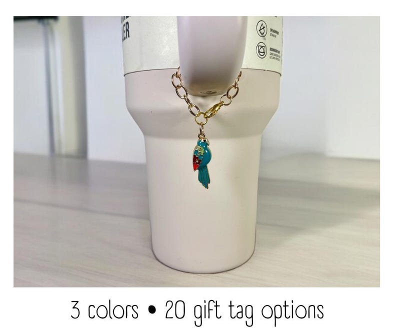 May include: A white ceramic mug with a gold chain and a blue and red bird charm. The mug has a black dot and the text "3 colors &bull; 20 gift tag options".