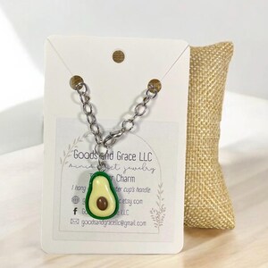 May include: A silver chain necklace with an avocado charm. The charm is green and yellow with a brown pit. The necklace is displayed on a white card with the text "Goods and Grace LLC" and "miniest jewelry".