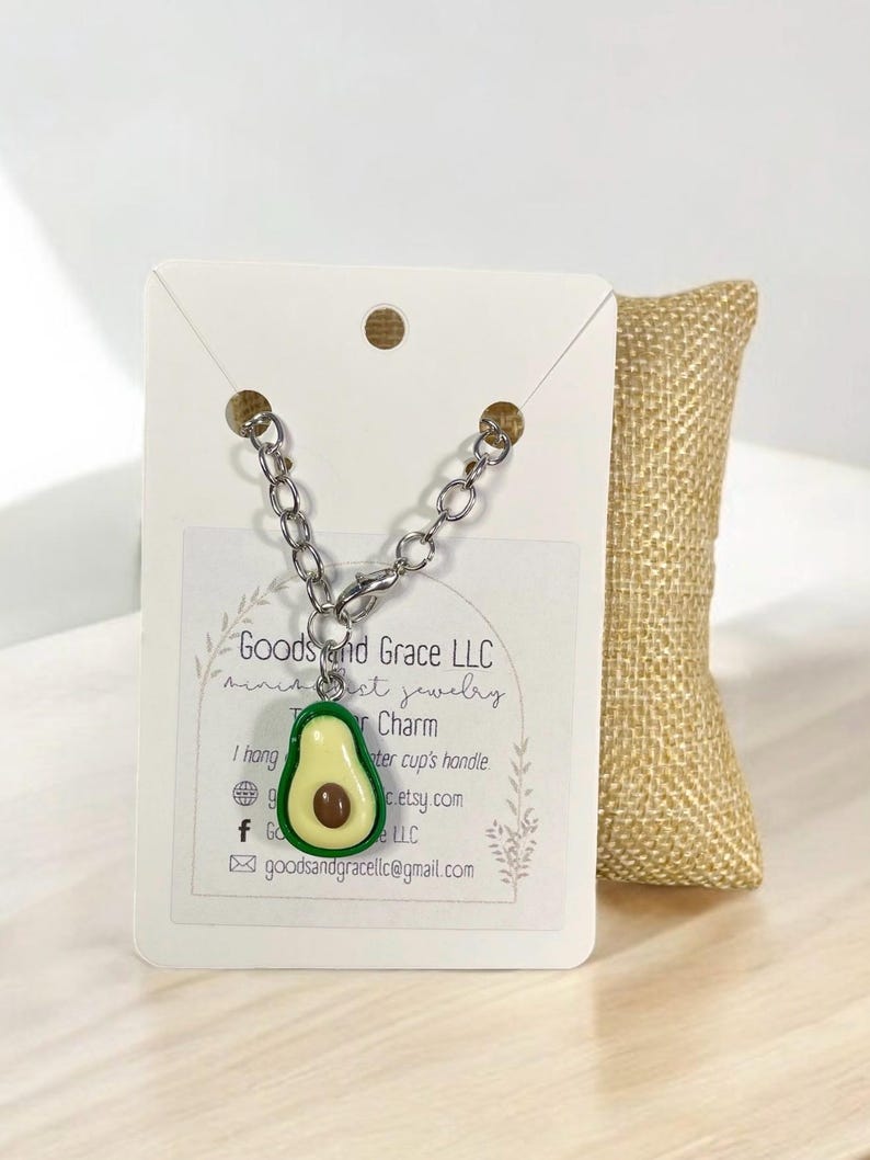 May include: A silver chain necklace with an avocado charm. The charm is green and yellow with a brown pit. The necklace is displayed on a white card with the text "Goods and Grace LLC" and "miniest jewelry".