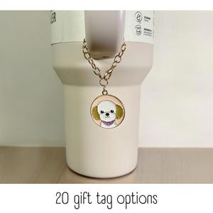 Può includere: Una catena dorata con un ciondolo rotondo che presenta un cane bianco dei cartoni animati con orecchie verdi e una bandana viola. Il ciondolo è appeso a una tazza bianca con il testo "20 opzioni di etichette regalo".