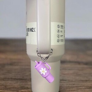 May include: A light gray stainless steel tumbler with a light blue lid and a handle. A purple charm with a white daisy is attached to the tumbler.