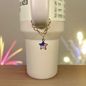 May include: A gold chain with a star-shaped charm featuring the American flag colors, red, white, and blue, hanging from a white travel mug.