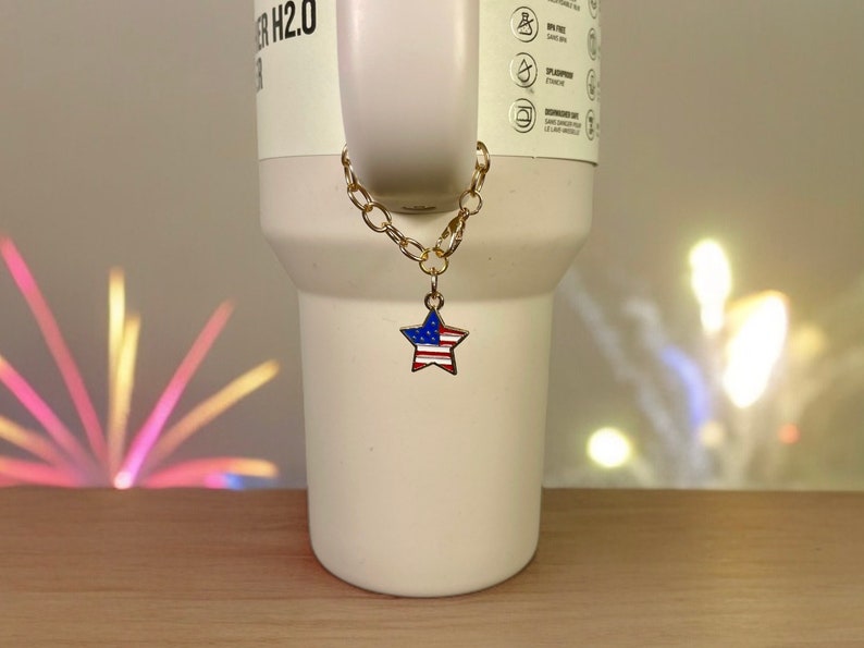 May include: A gold chain with a star-shaped charm featuring the American flag colors, red, white, and blue, hanging from a white travel mug.
