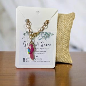 May include: A gold chain bracelet with a pink and green parrot charm. The charm is about 4.25 inches long. The bracelet is handmade and comes with a small burlap bag. The tag says "Goe & Grace Handmade Jewelry, Key Cup Charm, Size (-4.25"), $8, goodedgracelic, goodsandgracelic.etsy.com"