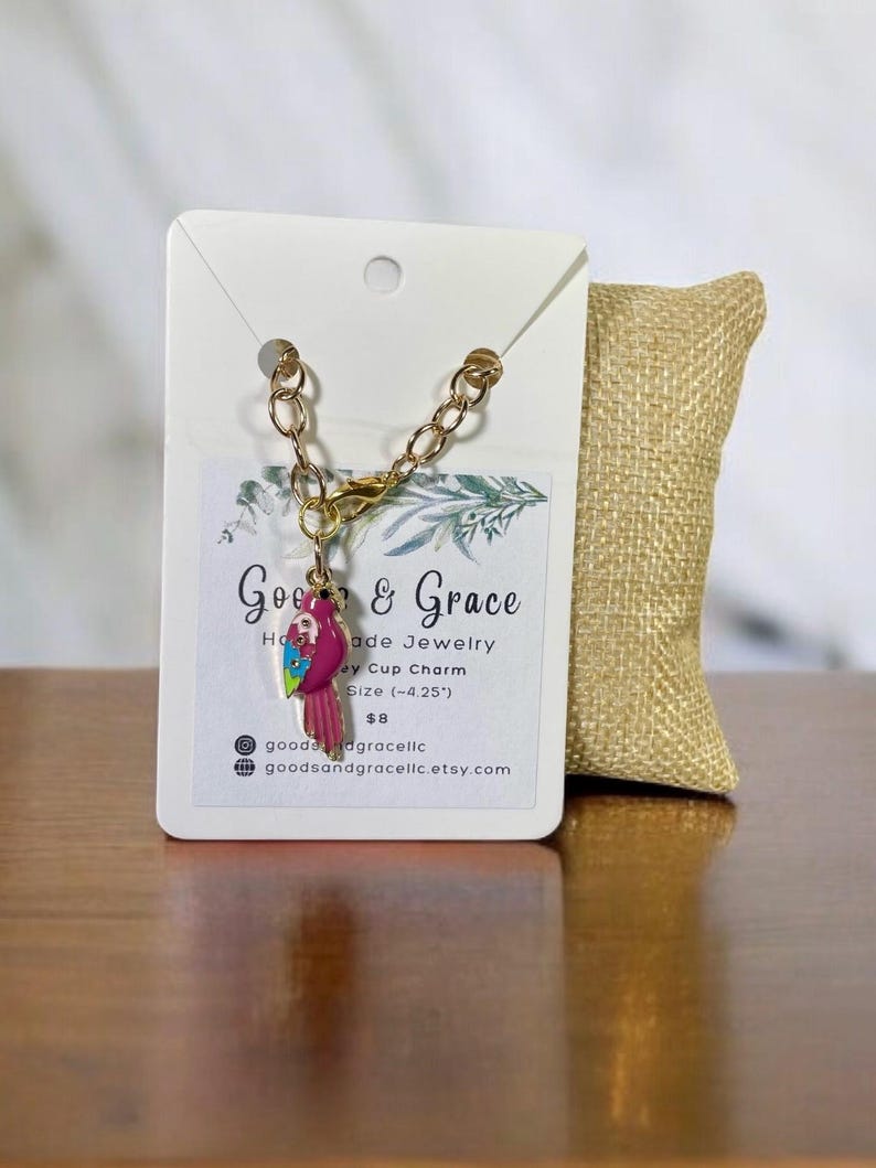 May include: A gold chain bracelet with a pink and green parrot charm. The charm is about 4.25 inches long. The bracelet is handmade and comes with a small burlap bag. The tag says "Goe & Grace Handmade Jewelry, Key Cup Charm, Size (-4.25"), $8, goodedgracelic, goodsandgracelic.etsy.com"