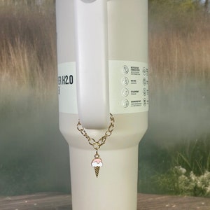 May include: A white stainless steel tumbler with a gold chain and ice cream charm. The tumbler has a blue lid and a handle. The tumbler has text that reads "H2.0".