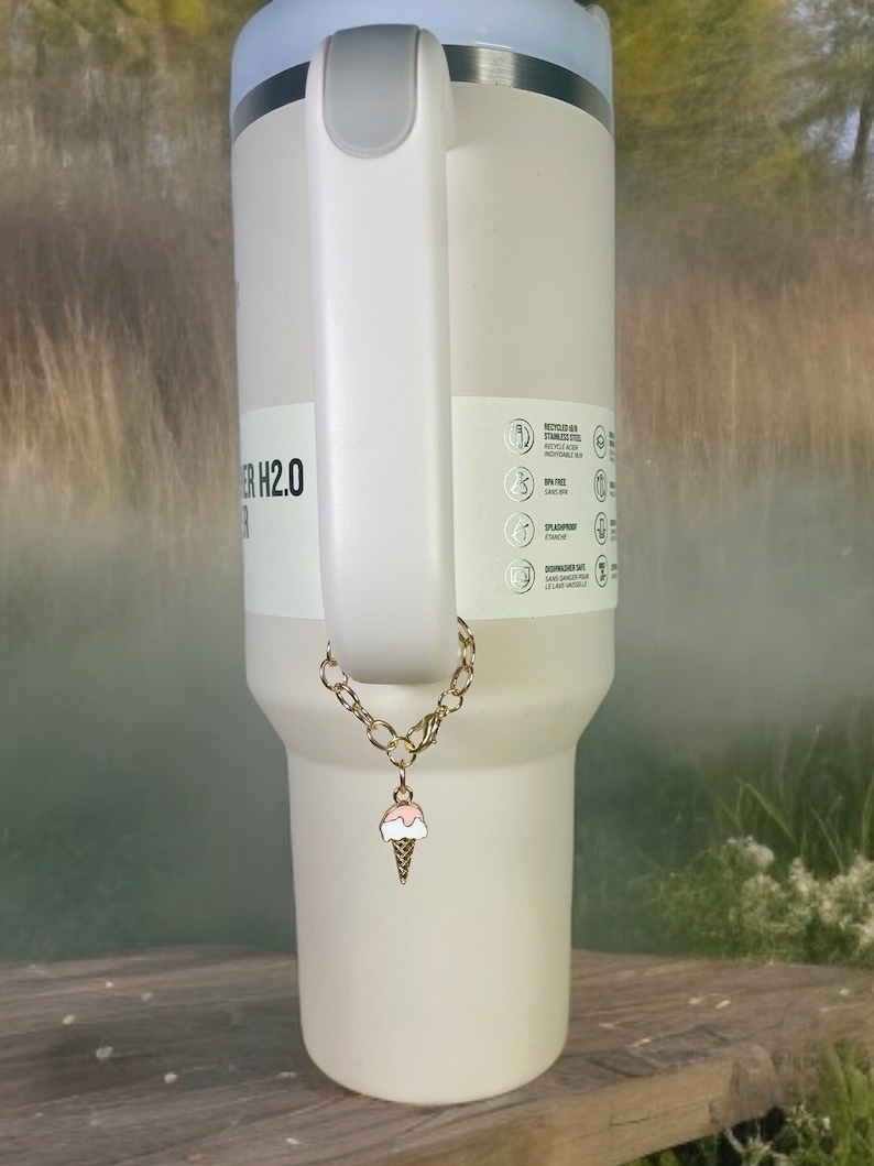 May include: A white stainless steel tumbler with a gold chain and ice cream charm. The tumbler has a blue lid and a handle. The tumbler has text that reads "H2.0".