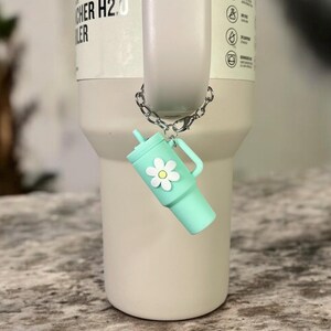 May include: A silver chain with a light blue and white charm attached to a stainless steel tumbler. The charm is shaped like a small tumbler with a white daisy on the side. The tumbler has the text "CHER H2.0 LER" on the side.