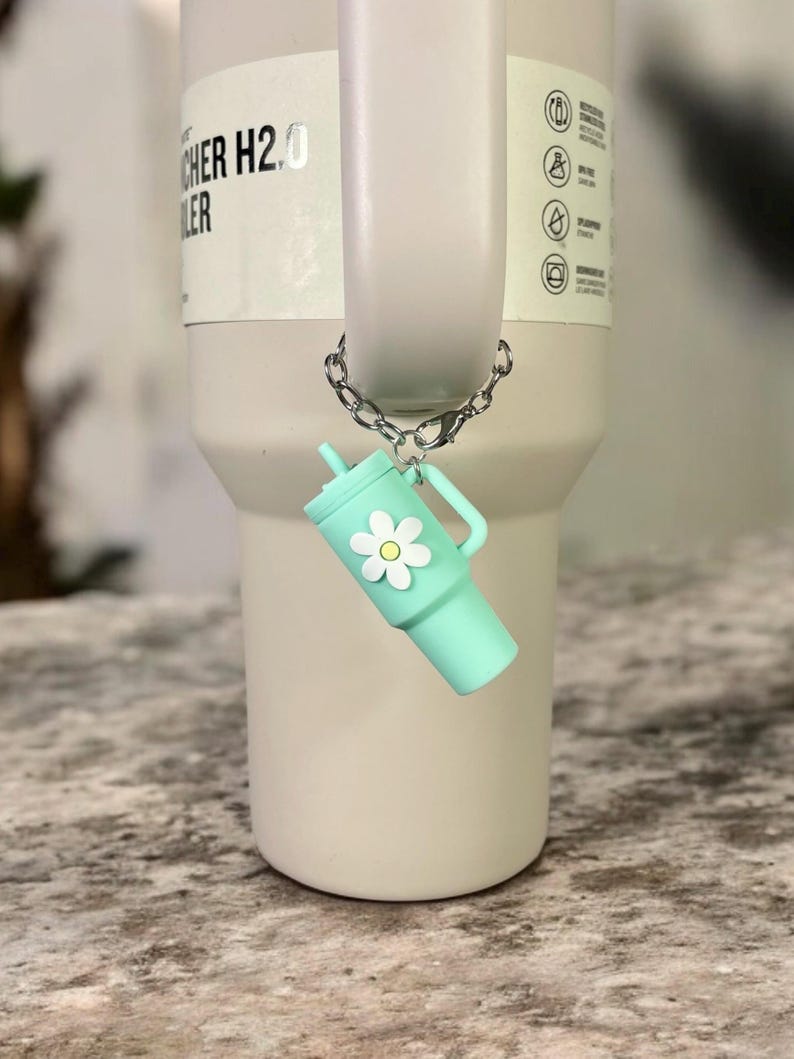 May include: A silver chain with a light blue and white charm attached to a stainless steel tumbler. The charm is shaped like a small tumbler with a white daisy on the side. The tumbler has the text "CHER H2.0 LER" on the side.