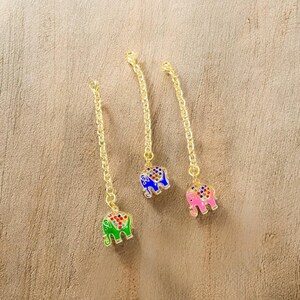 May include: Three gold-colored chain pendants, each with a colorful elephant charm. The elephants are green, blue, and pink, with intricate patterns and jewel-like accents. The background is a light brown, textured surface.