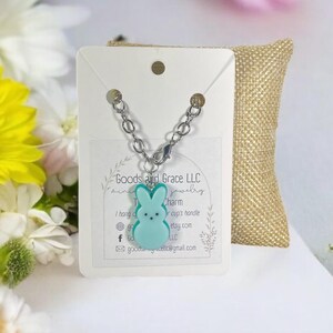 May include: A teal blue bunny charm on a silver chain. The charm is a cute, cartoon-style bunny with a white face and black eyes. The charm is hanging from a silver chain with a lobster clasp. The charm is on a white card with the text "Goods and Grace LLC Jewelry" and other information about the shop.