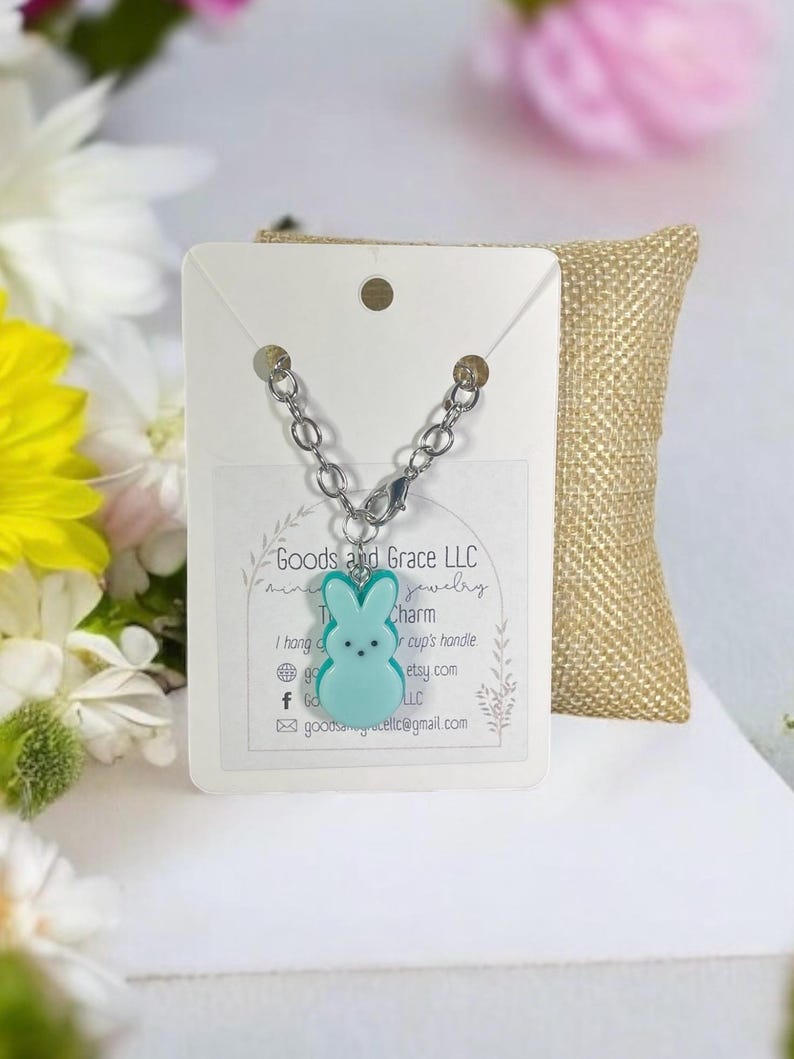 May include: A teal blue bunny charm on a silver chain. The charm is a cute, cartoon-style bunny with a white face and black eyes. The charm is hanging from a silver chain with a lobster clasp. The charm is on a white card with the text "Goods and Grace LLC Jewelry" and other information about the shop.