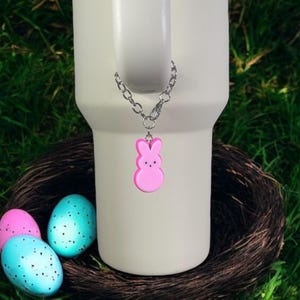 May include: A white travel mug with a pink bunny charm attached to a silver chain. The mug is sitting on a brown nest with three speckled Easter eggs.