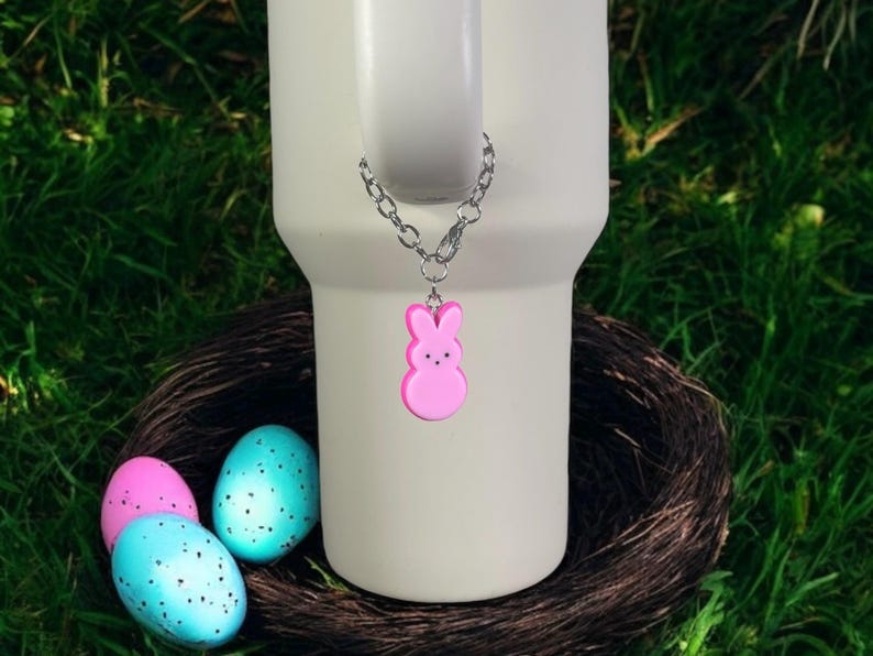 May include: A white travel mug with a pink bunny charm attached to a silver chain. The mug is sitting on a brown nest with three speckled Easter eggs.