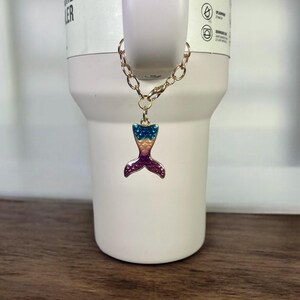 May include: A gold chain with a mermaid tail charm. The tail is made of metal and has a pink, blue, and yellow design.